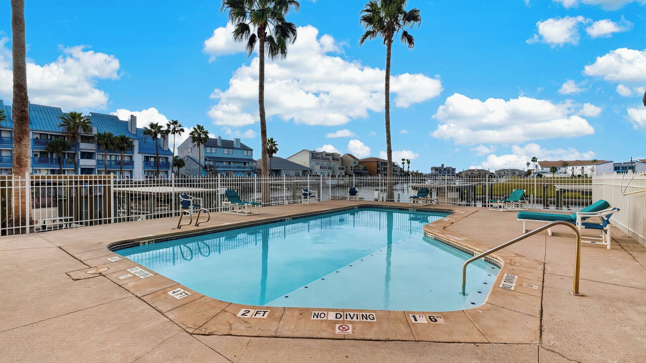A pool area with a diving board, palm trees, and a white fence.
