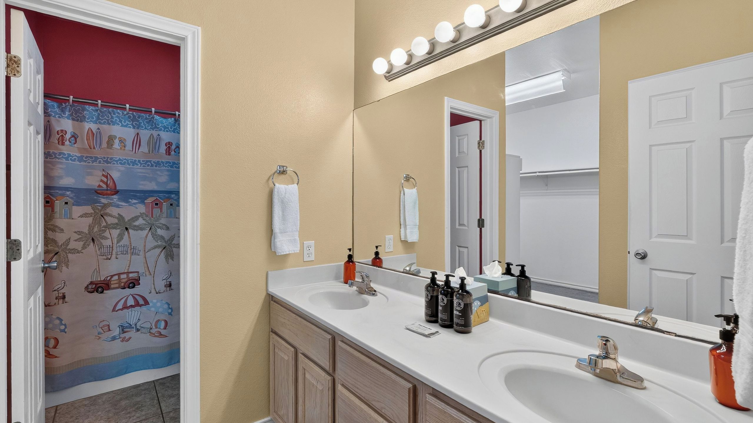 Un baño con lavamanos doble, tocador y espejo con luces. Hay un perchero y una puerta con un detalle rojo.