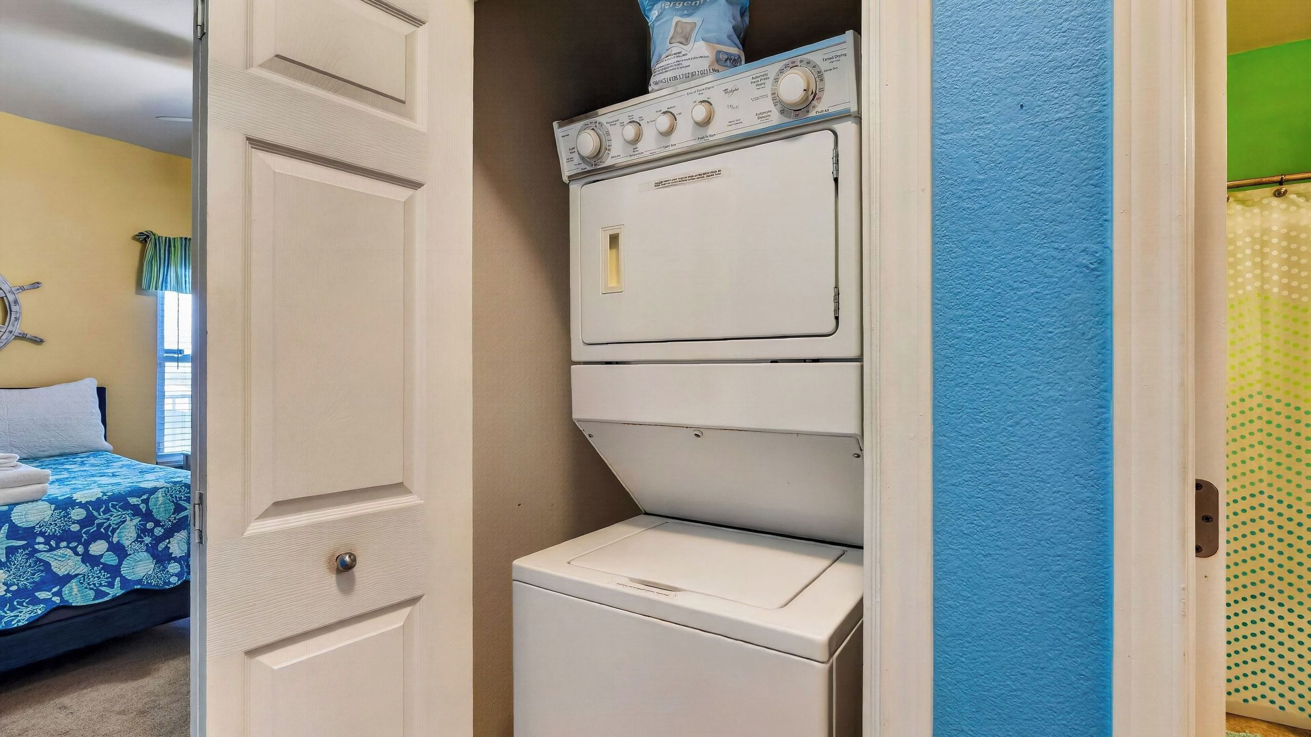 A laundry room with stacked washers and dryers, a bed with a blue and white bedspread, and a wall-mounted clock.