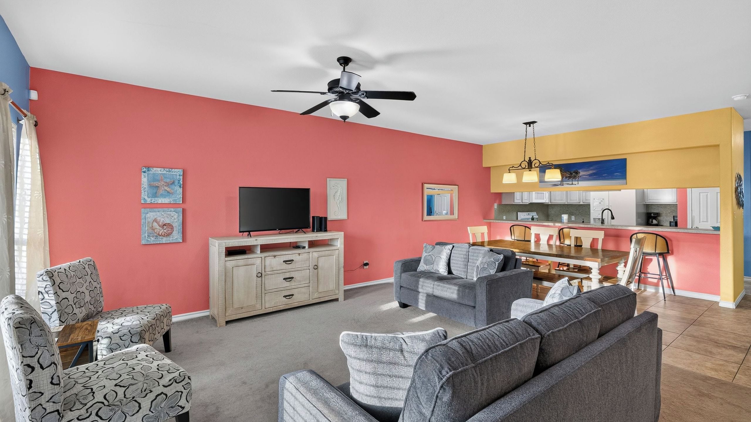 A living room with a flat-screen TV, a ceiling fan, and a dining area with a kitchen in the background.