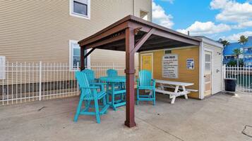 A covered outdoor seating area with turquoise chairs and a picnic table, adjacent to a building with a pool.
