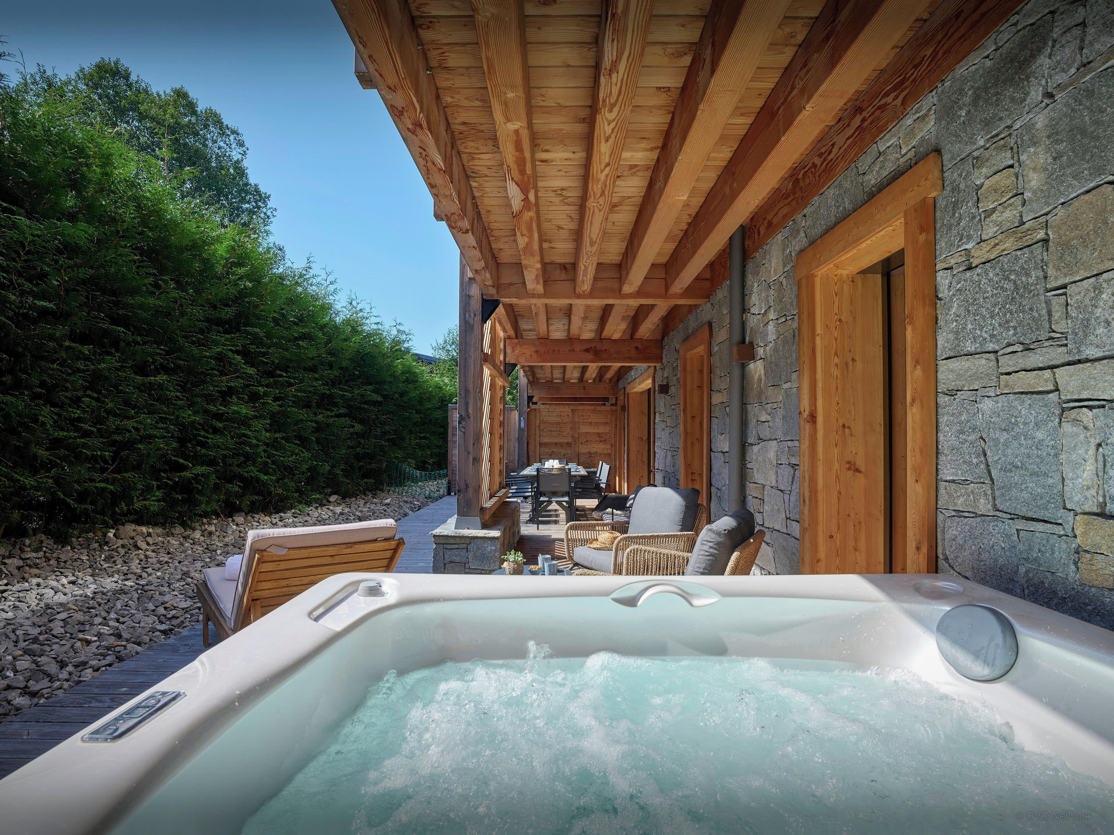 A hot tub with a wooden deck and stone walls, surrounded by outdoor furniture and a stone building.