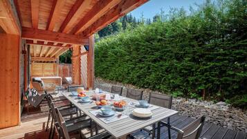 Een houten terras buiten met een eettafel, omringd door stoelen, en uitzicht op een dicht bos.