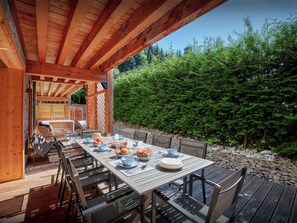 An outdoor wooden deck with a table set for dining, surrounded by chairs, and a view of a dense forest. - Lodge Ubuntu - modern apartment with a hot tub - OVO Network (Châtel)