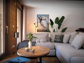A modern living room with a wooden floor, a round wooden coffee table, a grey sofa, a large plant, and a poster on the wall. - Lodge Ubuntu - modern apartment with a hot tub - OVO Network (Châtel)