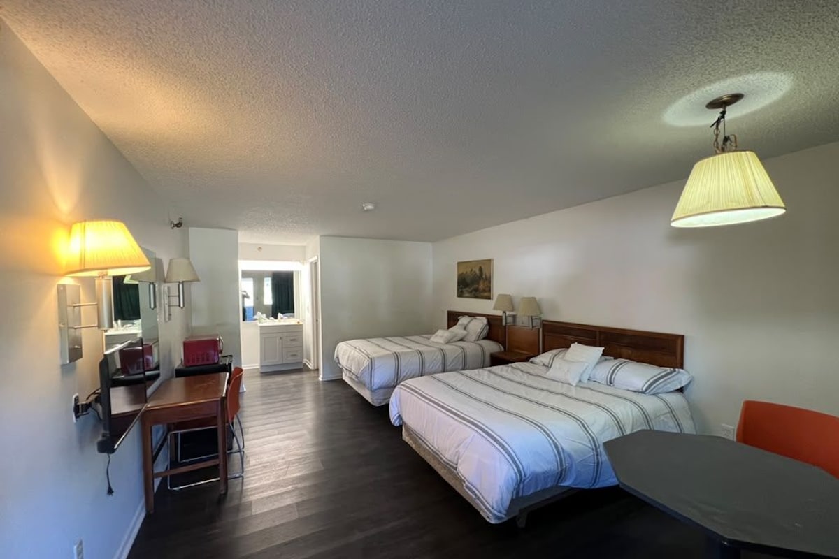 A hotel room with two beds, a dining table, a chair, a lamp, and a ceiling light fixture.
