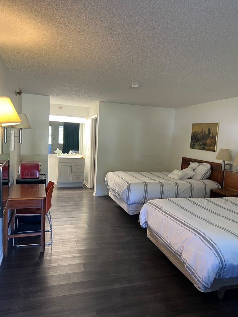 A hotel room with two beds, a desk, a chair, a small table, a lamp, and a painting on the wall.