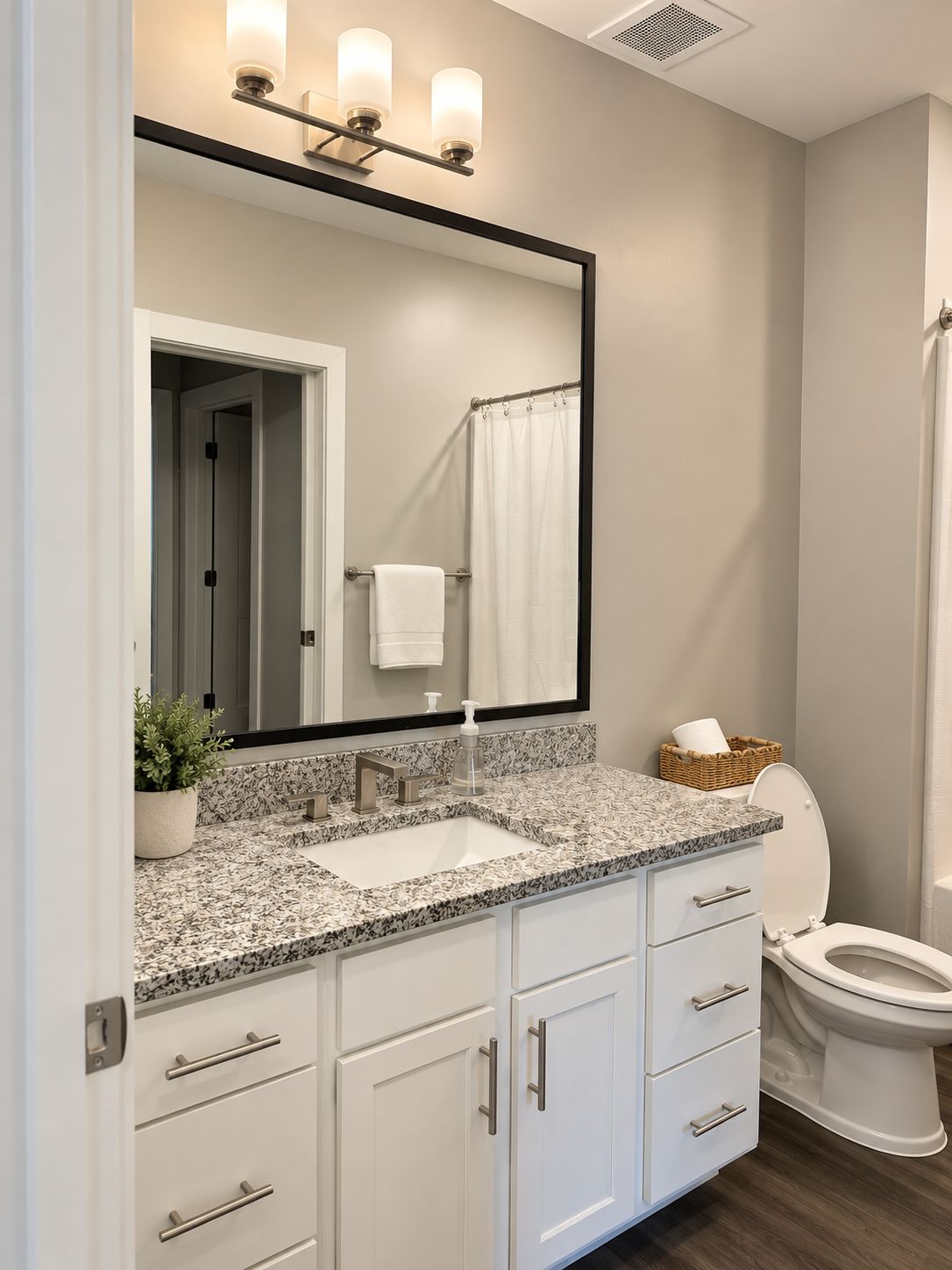 Un baño moderno con lavamanos de encimera de granito, armarios blancos, un espejo grande y un inodoro.
