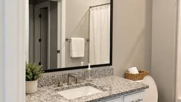 Un baño moderno con lavamanos de encimera de granito, armarios blancos, un espejo grande y un inodoro.