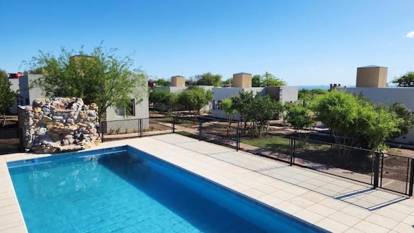 A swimming pool with a stone structure, surrounded by a fenced area with trees and buildings in the background.
