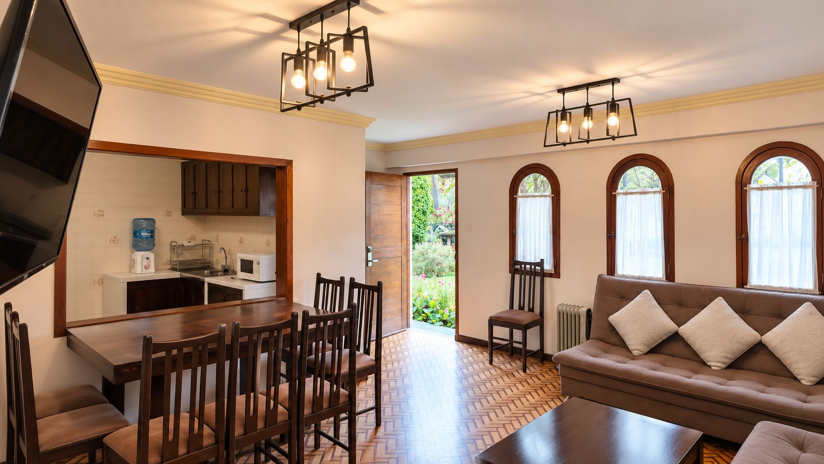 A living room with a brown sofa, wooden dining table, and chairs, a kitchen with white cabinets, and a large window with curtains.