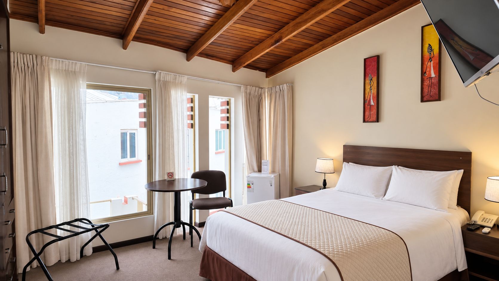 Quarto de hotel com cama, escrivaninha, cadeira, televisão e janela com cortinas.