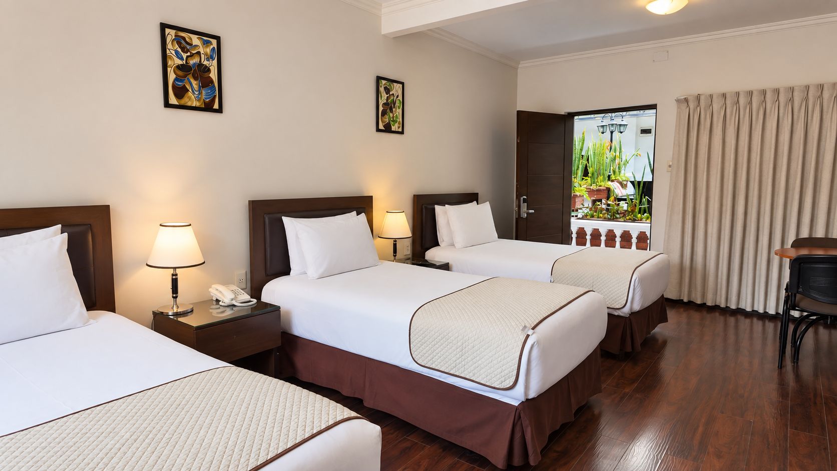 A hotel room with three beds, a desk, and a view of the garden through the door.