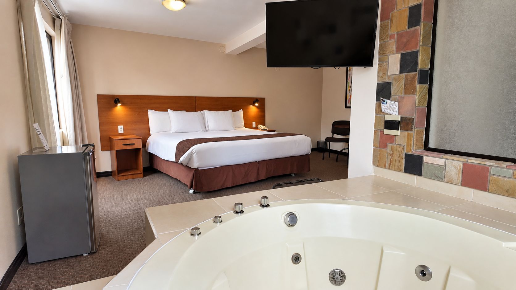 A hotel room with a large bed, a flat-screen TV, a minibar, and a bathtub.