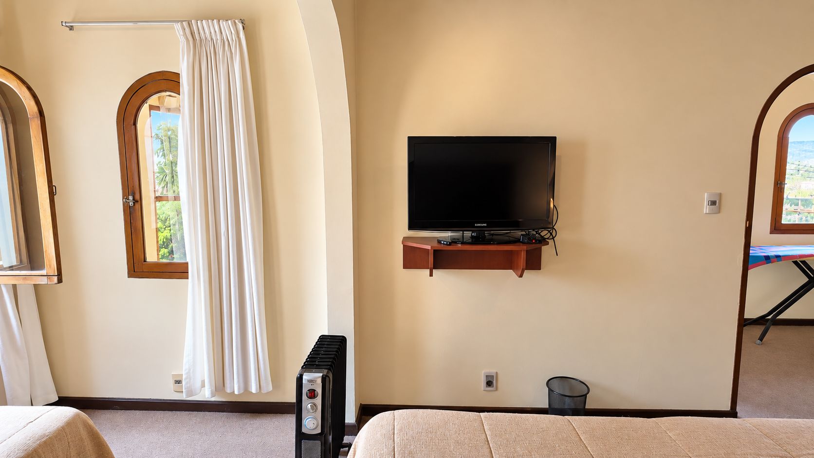 Una habitación con televisión montada en la pared, una ventana con cortinas, un radiador y una cama.