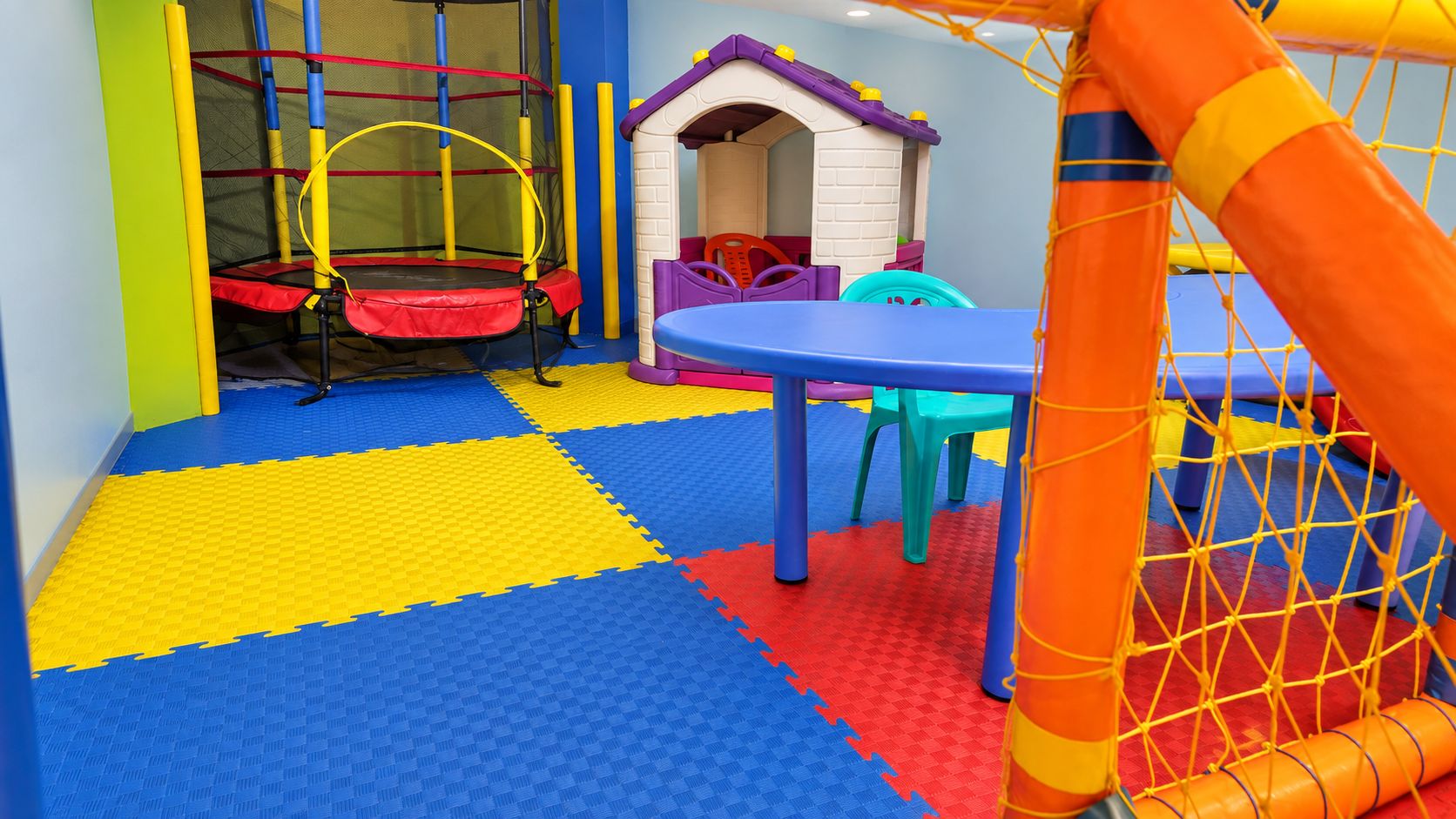 Zona de juegos con un trampolín, una casita pequeña y una estructura de escalada con red.