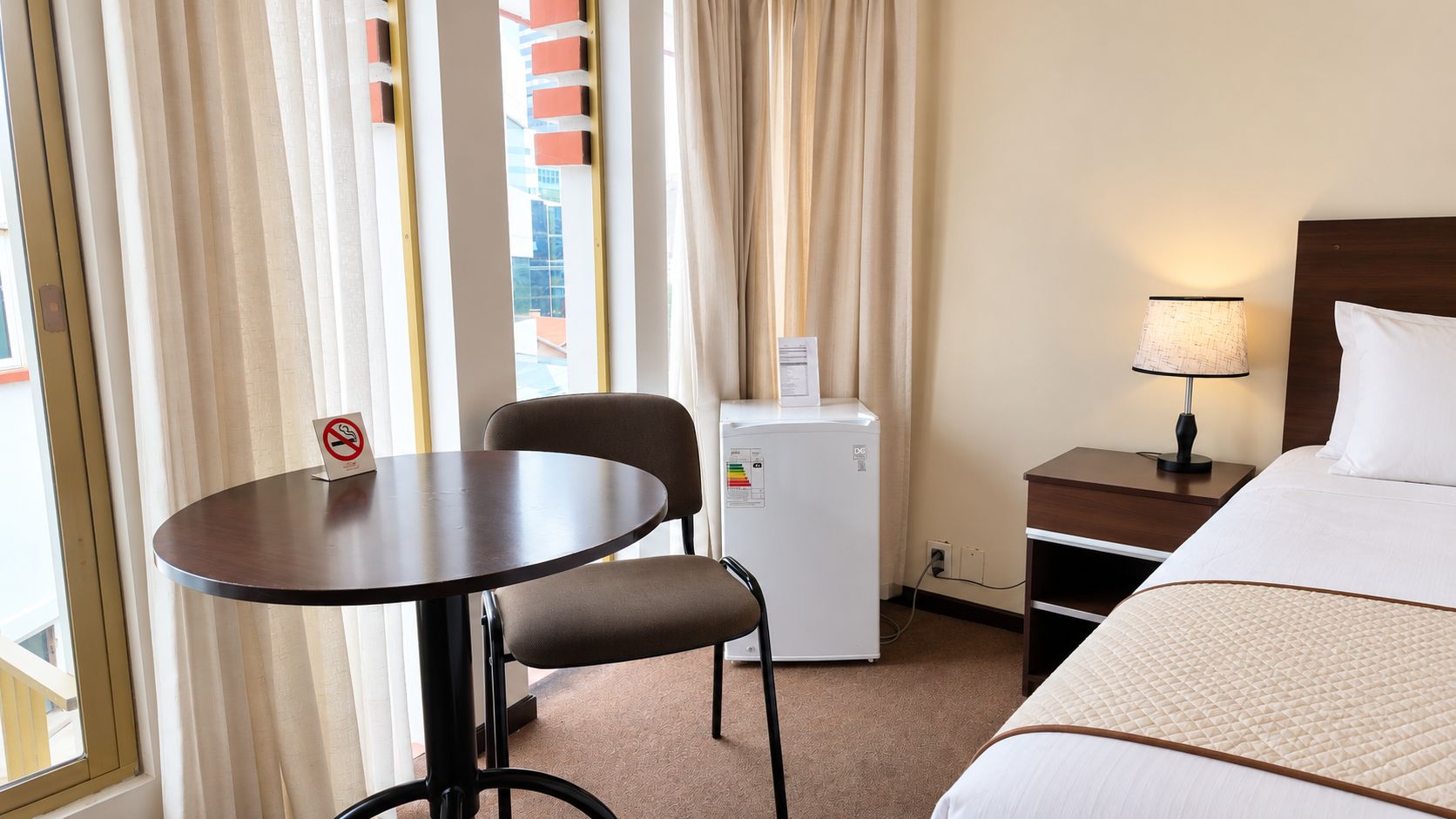 A hotel room with a bed, a table, a chair, a nightstand, a lamp, and a refrigerator.