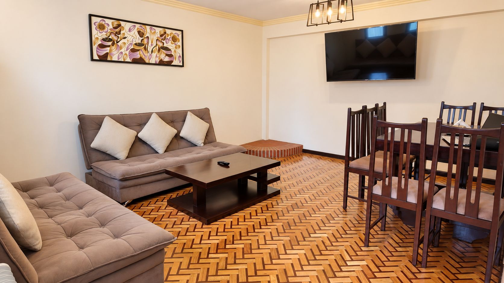 A living room with a sofa, a coffee table, a dining table with chairs, a television mounted on the wall, and a herringbone patterned floor.