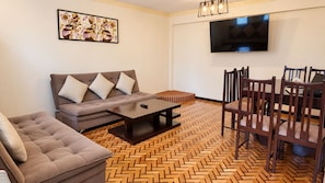 A living room with a sofa, a coffee table, a dining table with chairs, a television mounted on the wall, and a herringbone patterned floor. - Hotel Calacoto (La Paz)