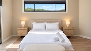 A hotel room with a large bed, two bedside tables with lamps, a window with a view of greenery, and a wooden floor. - The Ocean Dunes - Absolute Beachfront (The Entrance North)