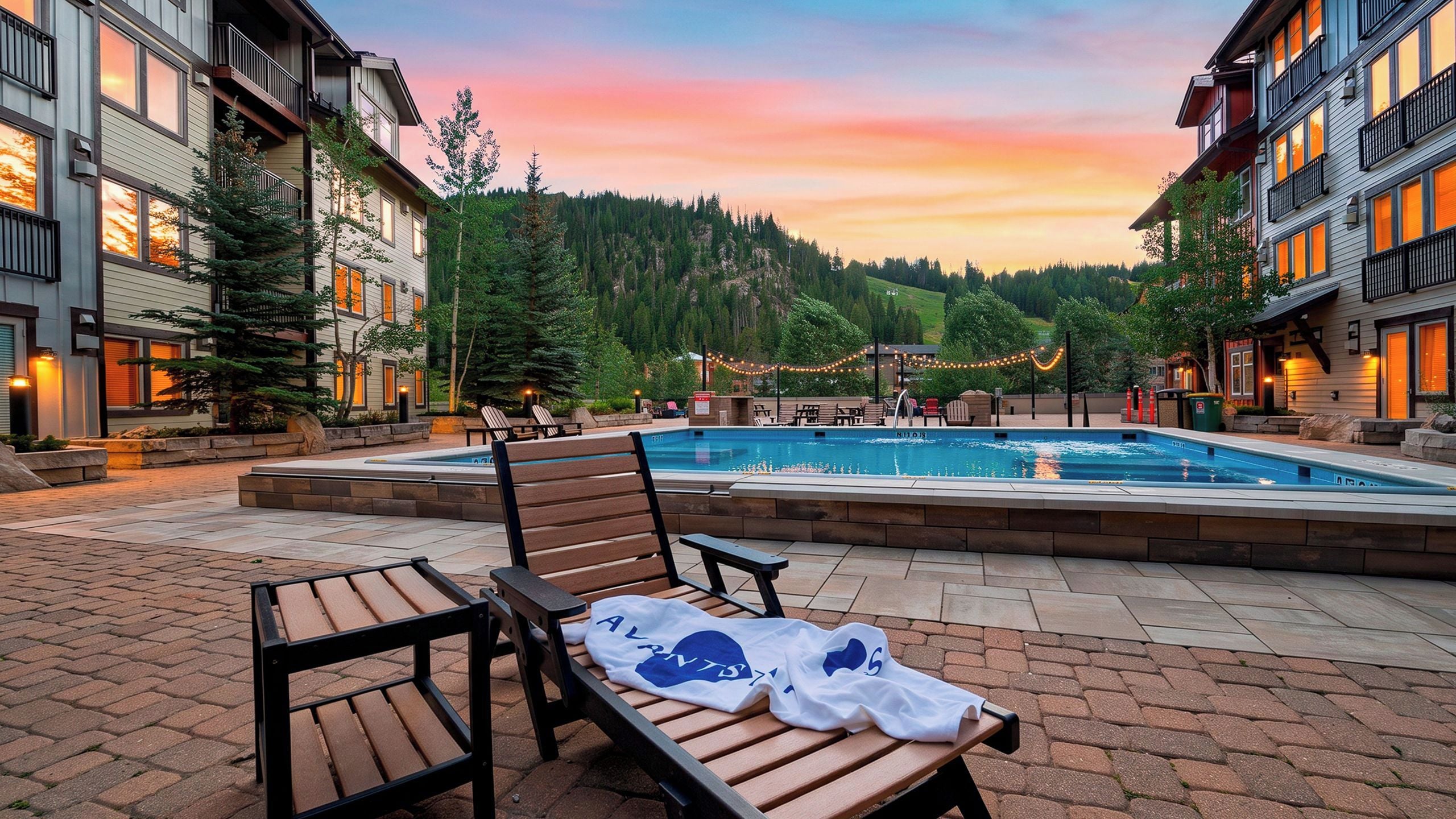A pool area with lounge chairs and a bench, surrounded by buildings and a forested hillside.