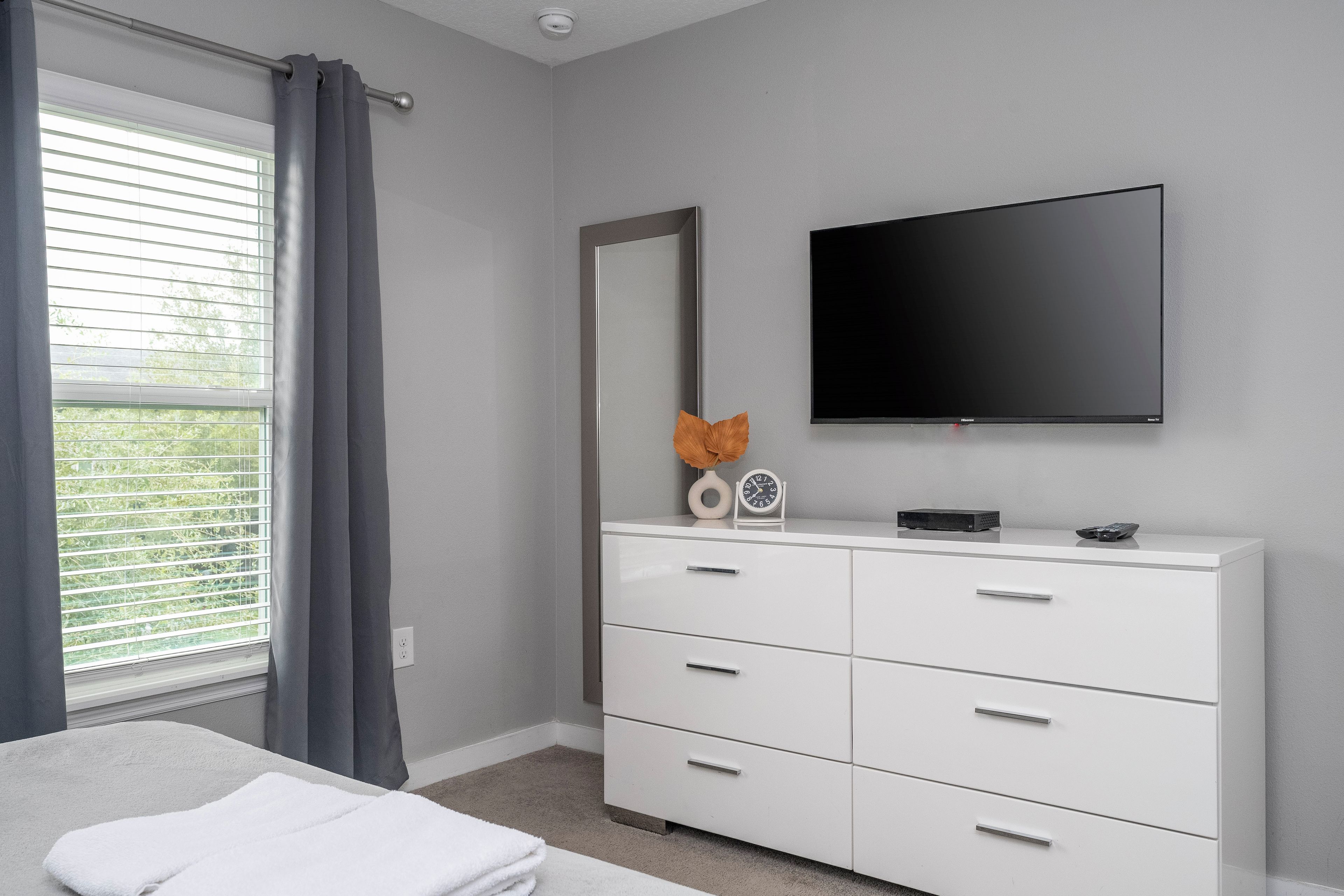 A bedroom with a bed, a dresser, a flat-screen TV, and a window with curtains.