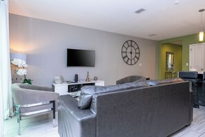 A modern living room with a grey sofa, a flat-screen TV, and a large wall clock. - 4BR Private Pool Near Disney & Supermarkets (Kissimmee)