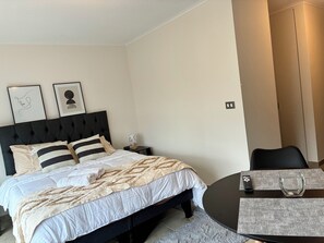 A hotel room with a bed, bedside table, lamp, framed artwork, and a round table with a remote control and glass. - Enjoy a modern and cozy studio apartment in Concepción. (Concepción)