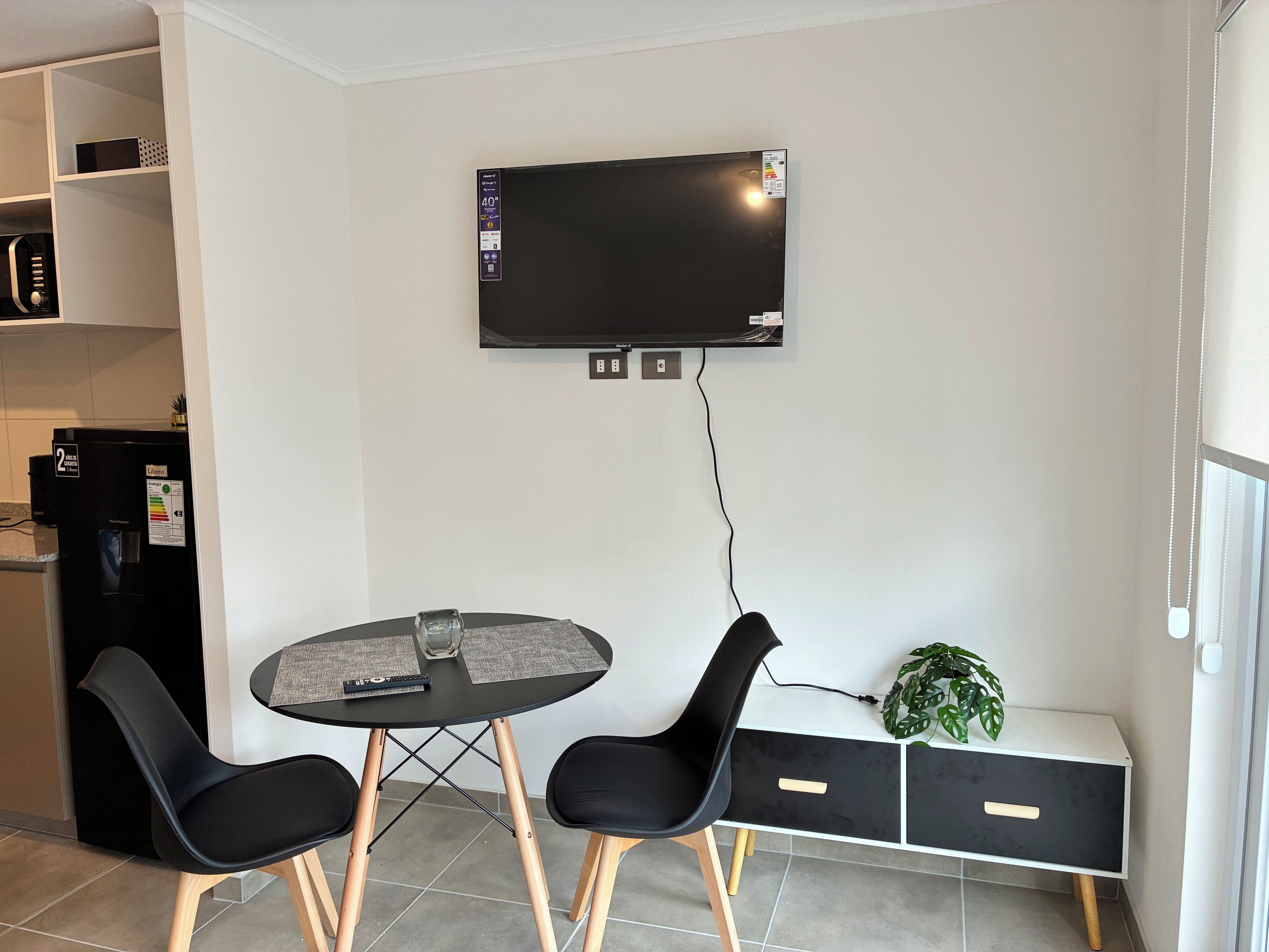 A modern kitchen with a built-in refrigerator, a flat-screen TV mounted on the wall, a round dining table with two chairs, and a small bench with a plant.