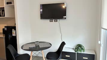 A modern kitchen with a built-in refrigerator, a flat-screen TV mounted on the wall, a round dining table with two chairs, and a small bench with a plant.
