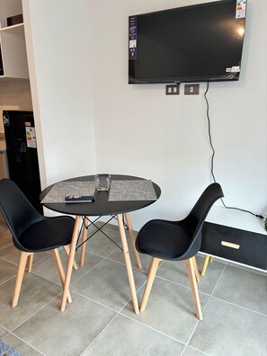 A room with a round table, two black chairs with wooden legs, a flat-screen TV mounted on the wall, and a small refrigerator in the corner. - Enjoy a modern and cozy studio apartment in Concepción. (Concepción)