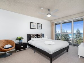 Ein modernes Schlafzimmer mit einem Bett, einem Nachttisch, einem Sessel und einem großen Fenster mit Blick auf Bäume.