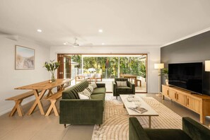 Ein geräumiges Wohnzimmer mit einer Couch, einem Couchtisch und einem Fernseher, verbunden mit einem Balkon mit Blick auf Grünflächen.