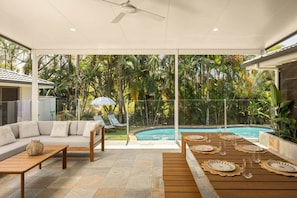 Ein moderner Außenbereich mit Pool, einer Sitzecke, einem Esstisch gedeckt mit Tellern und Gläsern sowie einem Deckenventilator.