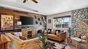 A modern living room with a flat-screen TV, wooden furniture, and a stone wall. - Rawhide Ranch by Avantstay Hot Tub + Fenced Yard + Grill Mountain Views + Near Park (Pioneertown)