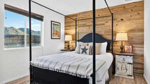 A four-poster bed with a view of mountains through a large window, a wooden wall, and a nightstand with a lamp. - Rawhide Ranch by Avantstay Hot Tub + Fenced Yard + Grill Mountain Views + Near Park (Pioneertown)
