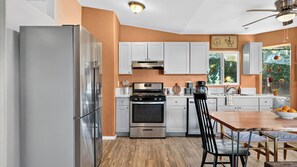 A modern kitchen with stainless steel appliances, white cabinets, and a dining area with a wooden table and chairs. - Rawhide Ranch by Avantstay Hot Tub + Fenced Yard + Grill Mountain Views + Near Park (Pioneertown)