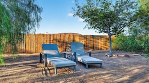 Two lounge chairs with a small table and a pitcher of water, set against a wooden fence and surrounded by trees. - Rawhide Ranch by Avantstay Hot Tub + Fenced Yard + Grill Mountain Views + Near Park (Pioneertown)