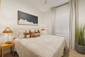Een net opgemaakt bed met een gewatteerde spreideken, twee bedlampjes, een aan de muur hangend schilderij en een raam met zonwering.
