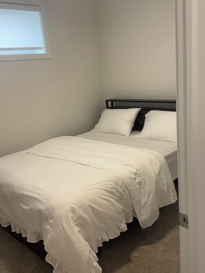 Un lit simple avec une literie blanche, dans une pièce dont le mur est uni et qui possède une fenêtre.