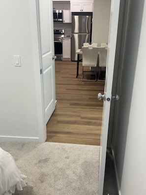 A hallway leading to a kitchen with white cabinets, stainless steel appliances, and a dining area with a white table and chairs. - Cozy 1 Bedroom Legal Basement Suite, Shops, transit close by. 15min. to Airport, (Edmonton)
