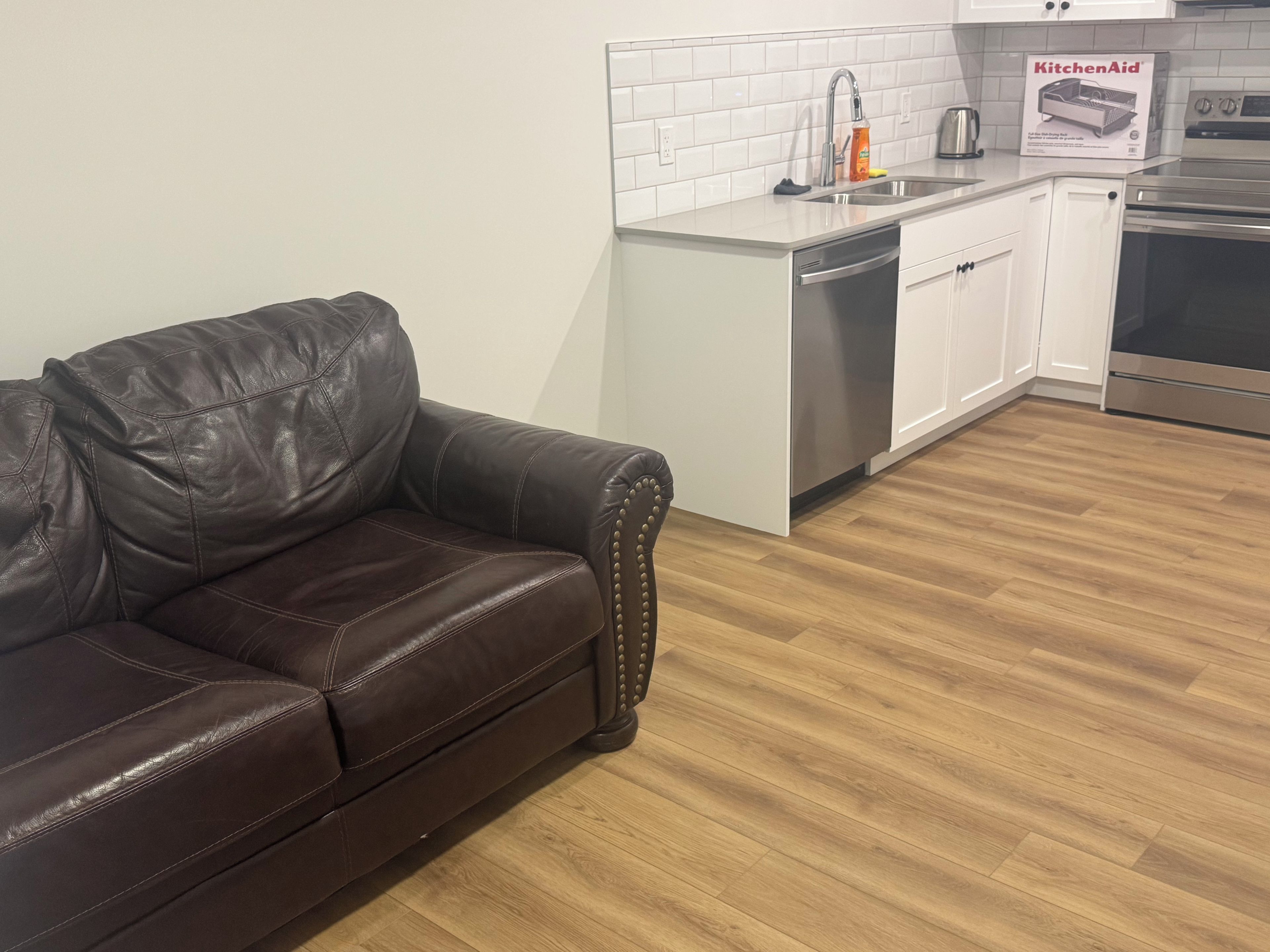 A modern kitchen with white cabinetry, stainless steel appliances, and a leather sofa in the living area. - Cozy 1 Bedroom Legal Basement Suite, Shops, transit close by. 15min. to Airport, (Edmonton)