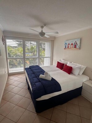 Ein Schlafzimmer mit Bett, Deckenventilator, Fenster mit Jalousien und einem Bild an der Wand.