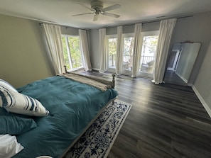 Ein Schlafzimmer mit einem Bett, einem Deckenventilator und Blick ins Freie durch große Fenster.