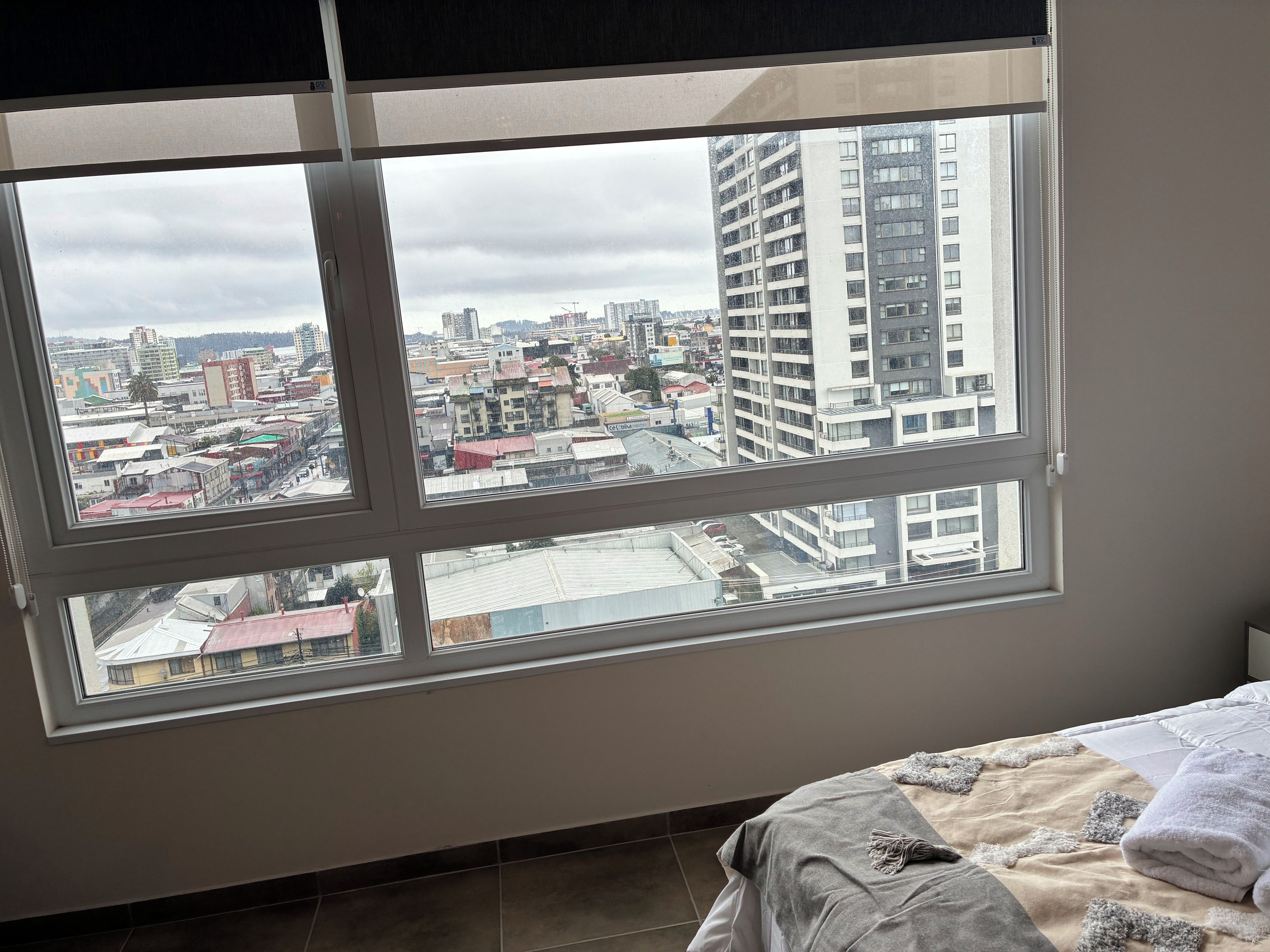 Una habitación con un ventanal que ofrece vistas a un paisaje urbano, una cama con una manta y un radiador fijado a la pared.