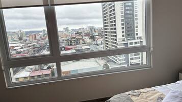 Una habitación con un ventanal que ofrece vistas a un paisaje urbano, una cama con una manta y un radiador fijado a la pared.
