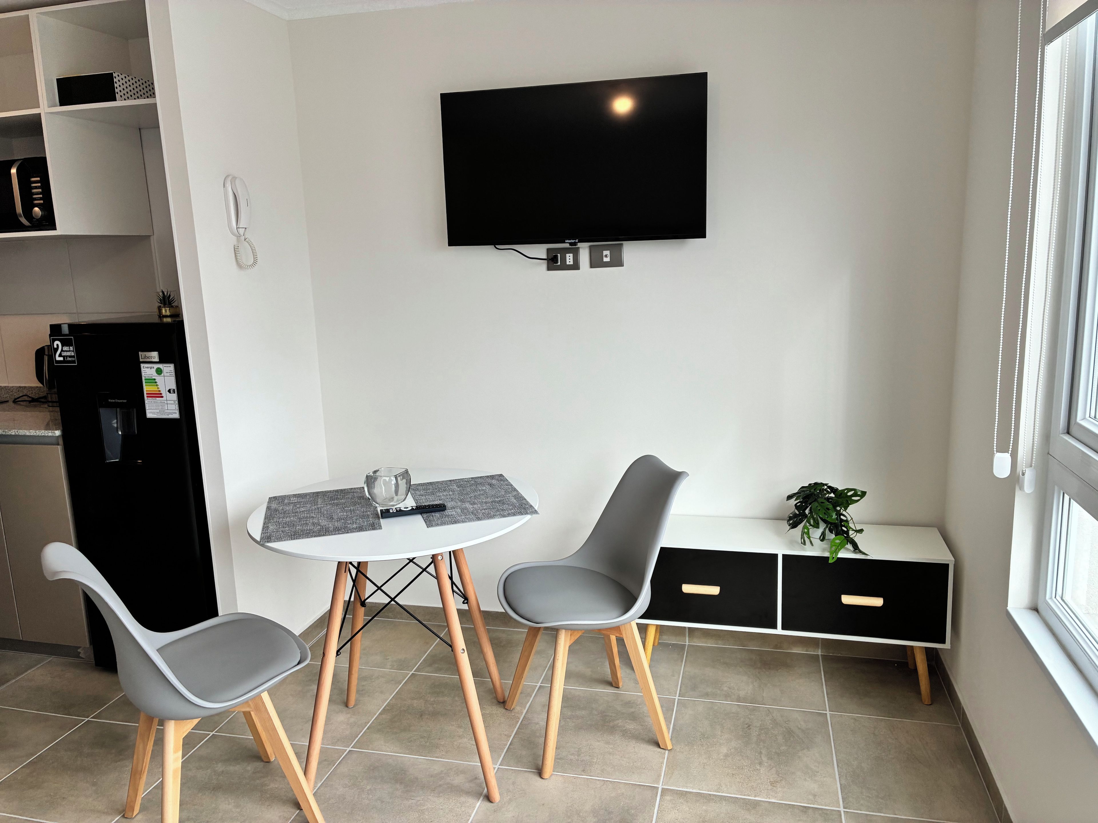 A modern kitchen with a dining area, a flat-screen TV mounted on the wall, a water dispenser, and a small plant.