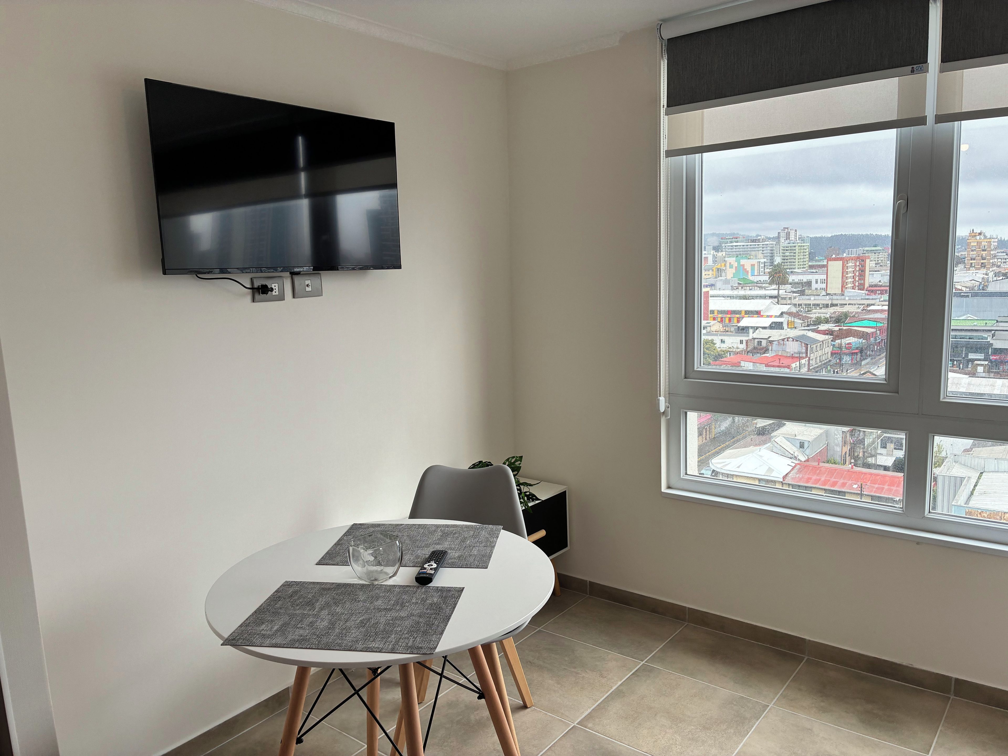 A small room with a TV mounted on the wall, a dining table with two chairs, a window with a city view, and a small cabinet.