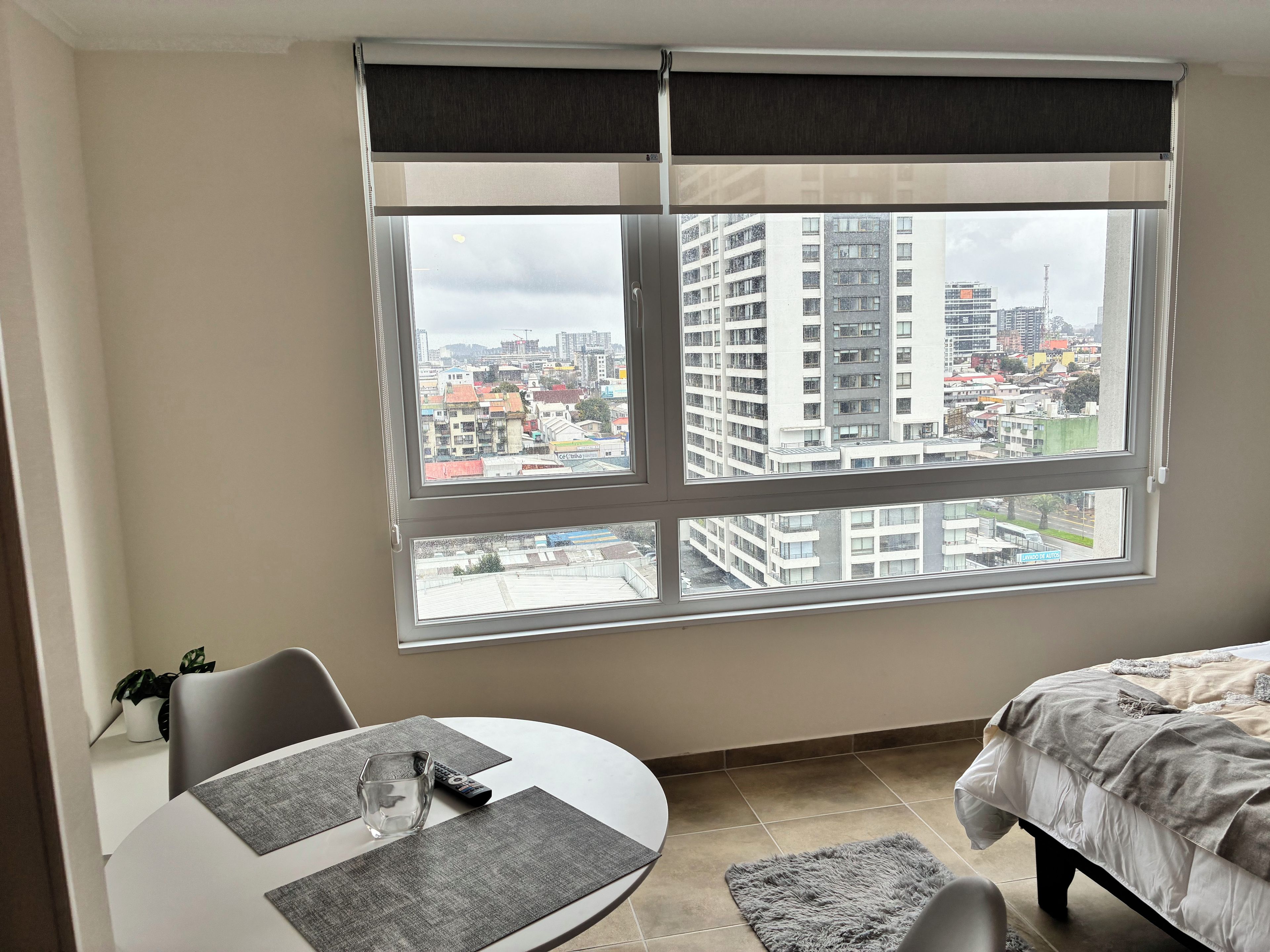 A room with a large window, a bed, a dining table with chairs, and a remote control on the table.