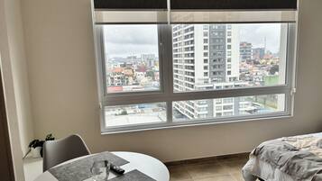 A room with a large window, a bed, a dining table with chairs, and a remote control on the table.